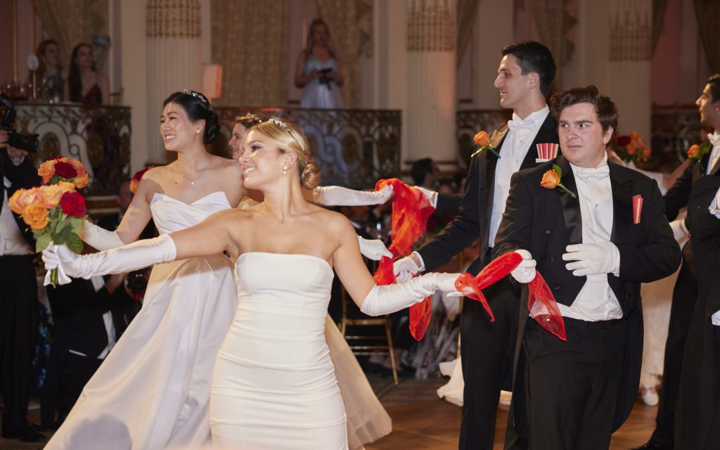 Debutantes and escorts wearing white dresses holding red fans line up to enter the hall